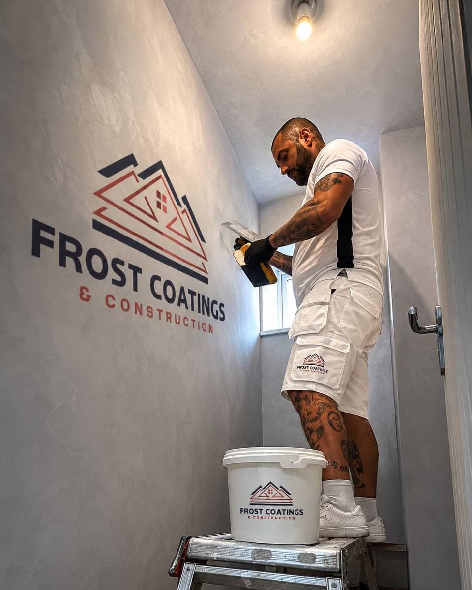 A polished plaster wall being worked by hand with a trowel, the Frost Coatings and Construction mark on the finished wall beside it
